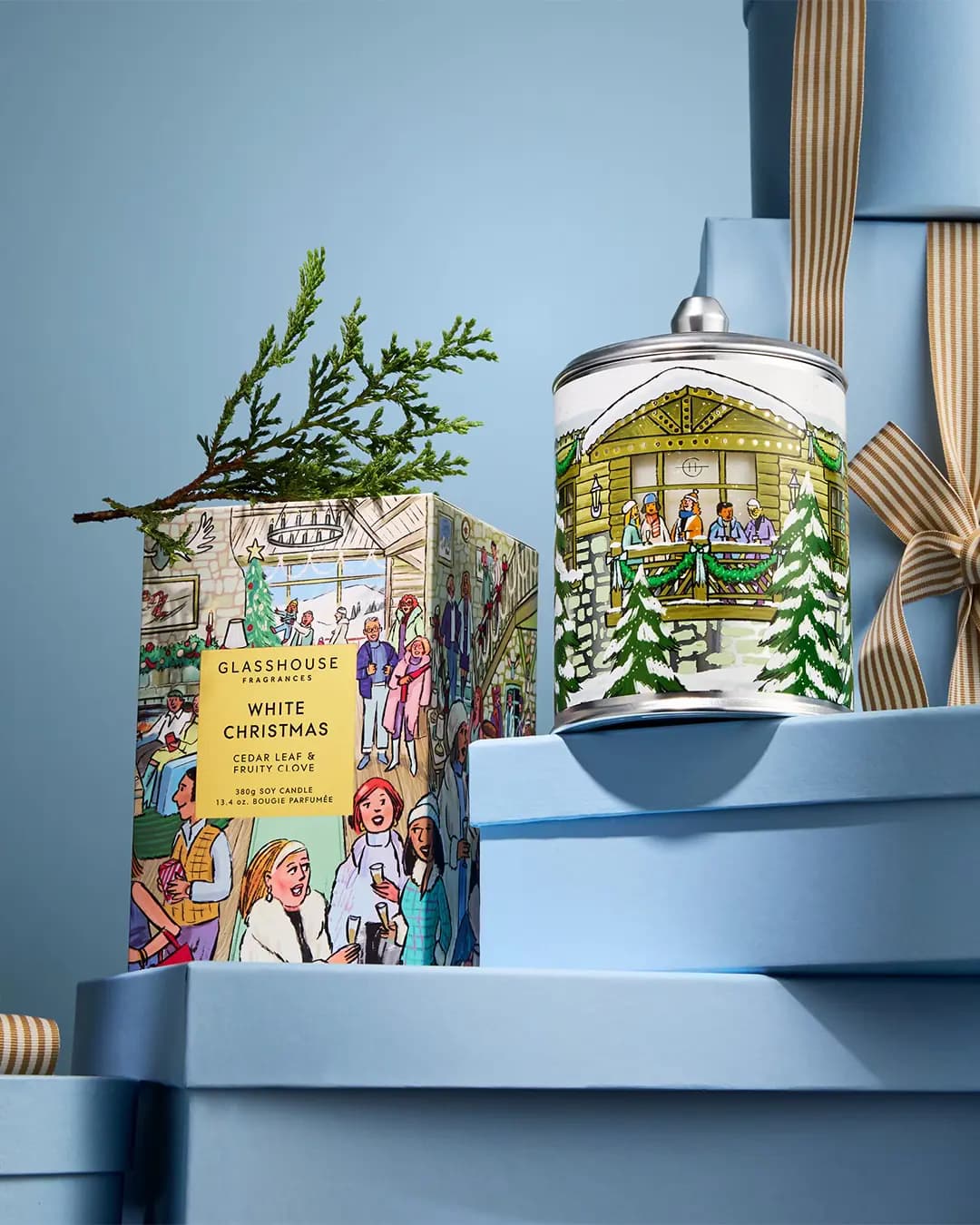 Glasshouse White Christmas silver-lidded tin with a snowy ski chalet next to its box topped with a cedar sprig on blue boxes