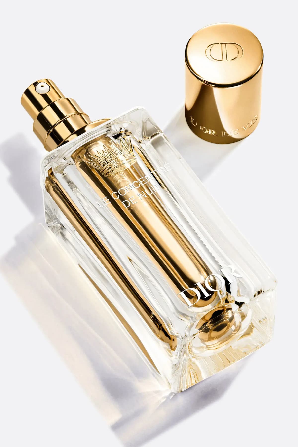 Uncapped bottle lying diagonally with gold pump and gold CD-embossed cap beside it on a white surface