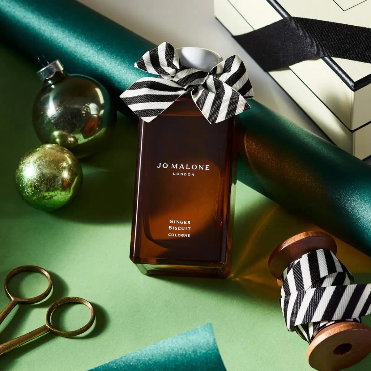 Amber cologne bottle with striped bow styled on green paper beside baubles, ribbon spools and brass scissors.