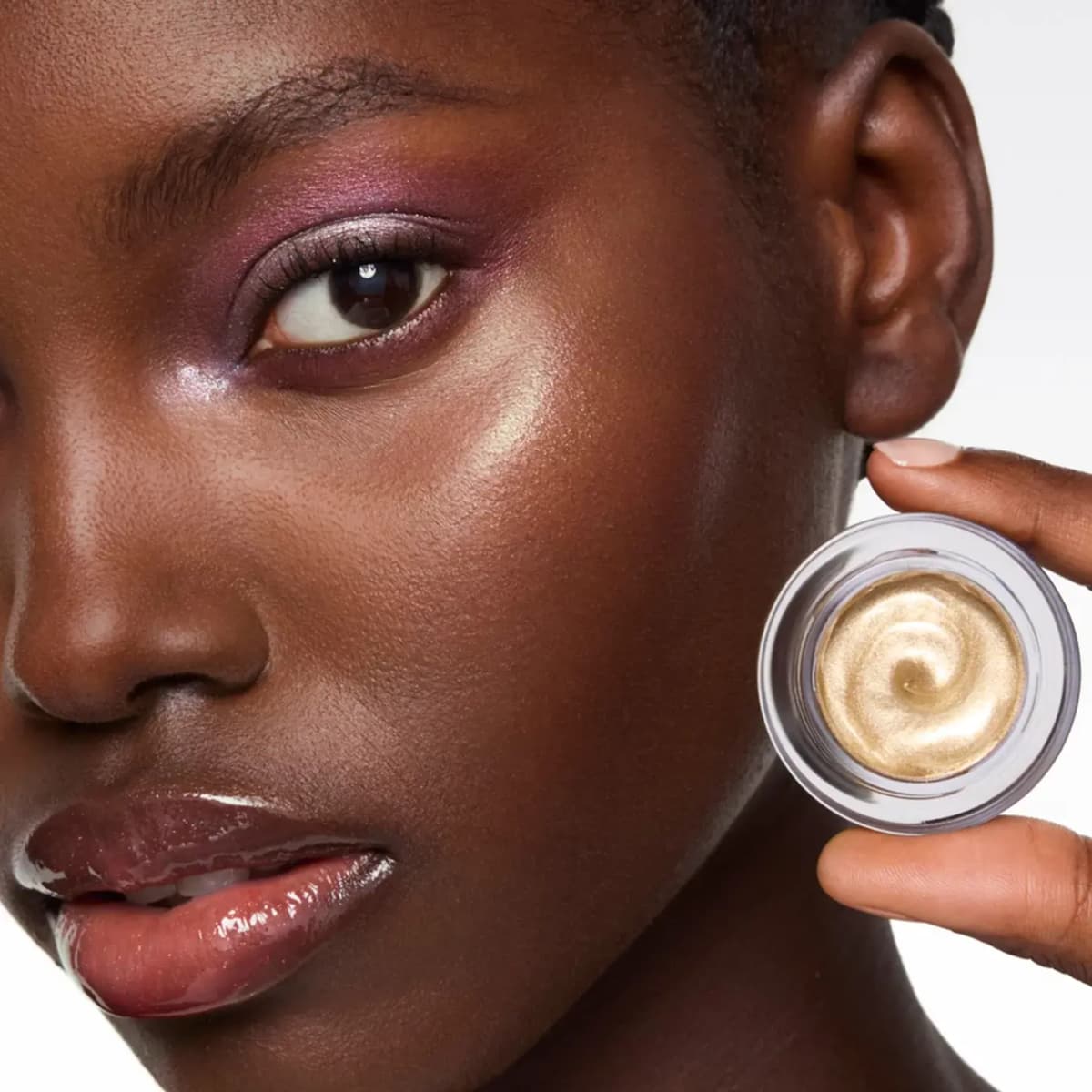 Model with plum eyeshadow and glossy lips holds an open pot of swirled gold highlighter near her glowing cheek
