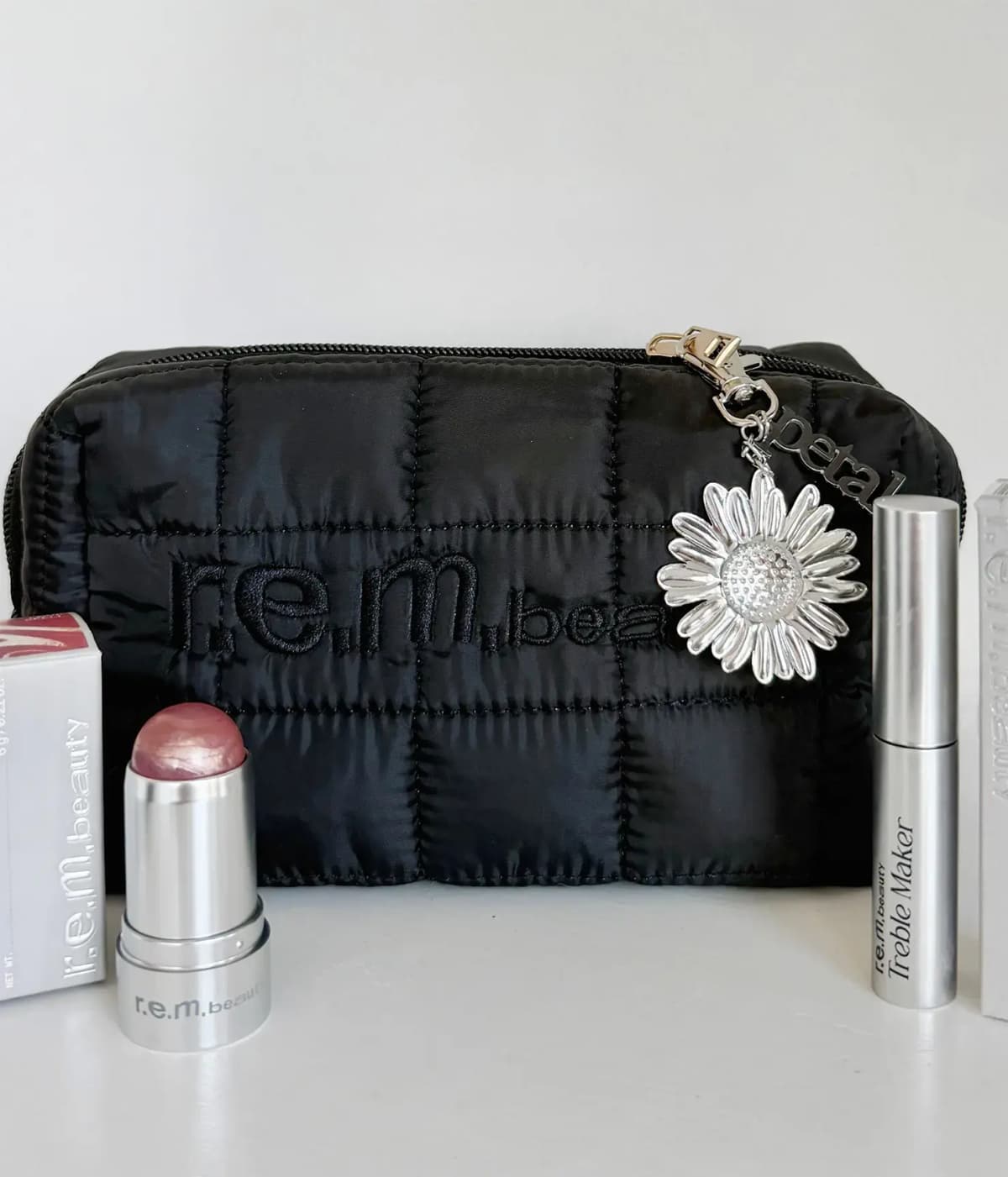 Quilted bag showing r.e.m. beauty embroidery, daisy keychain clipped to zipper, mauve blush stick and mascara with boxes