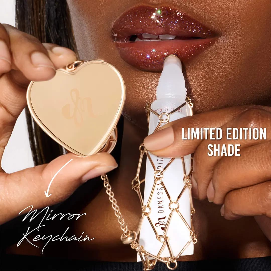 Model with glittery brown lips holds the caged white gloss tube to the lips beside the gold heart mirror keychain