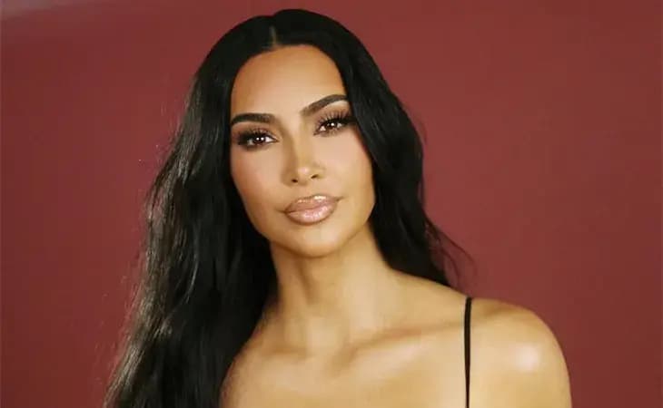 Kim Kardashian smiling softly in a black strappy top against a deep red background, with soft waves and nude lipstick.