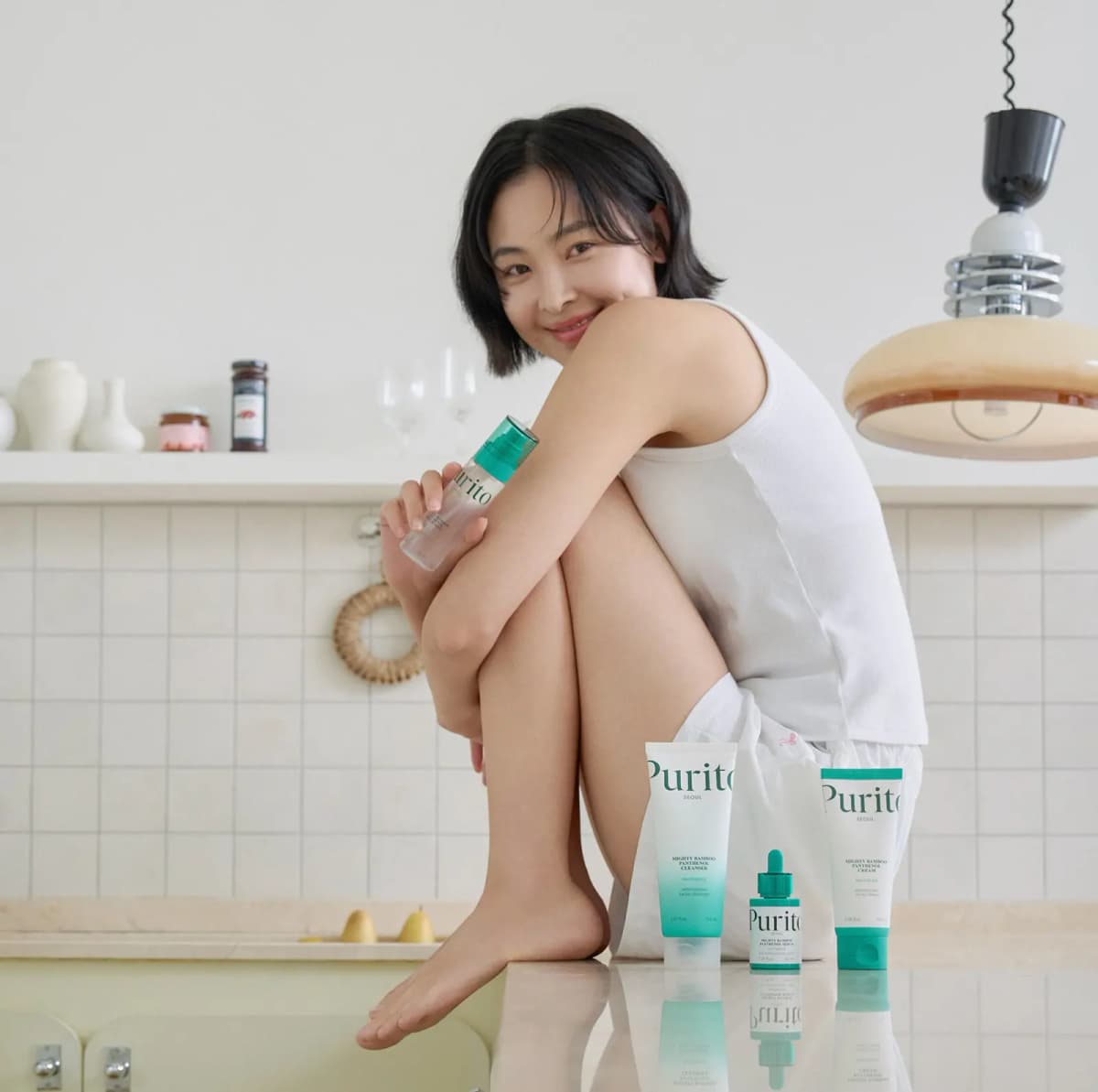 Smiling woman in a white tank top holds the cream mist on a kitchen counter beside Purito cleanser, serum and cream.