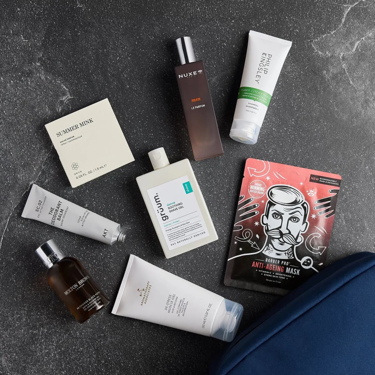 Flat lay of eight grooming products scattered on dark speckled stone beside the corner of a navy zip bag