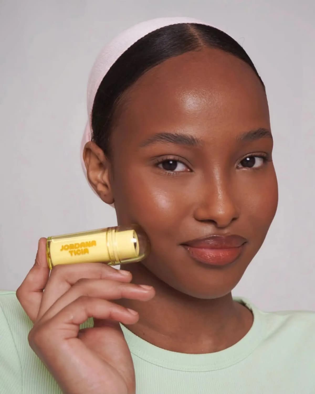 Model in a pink headband and mint top holds a capped Jordana Ticia stick to her glowing cheek