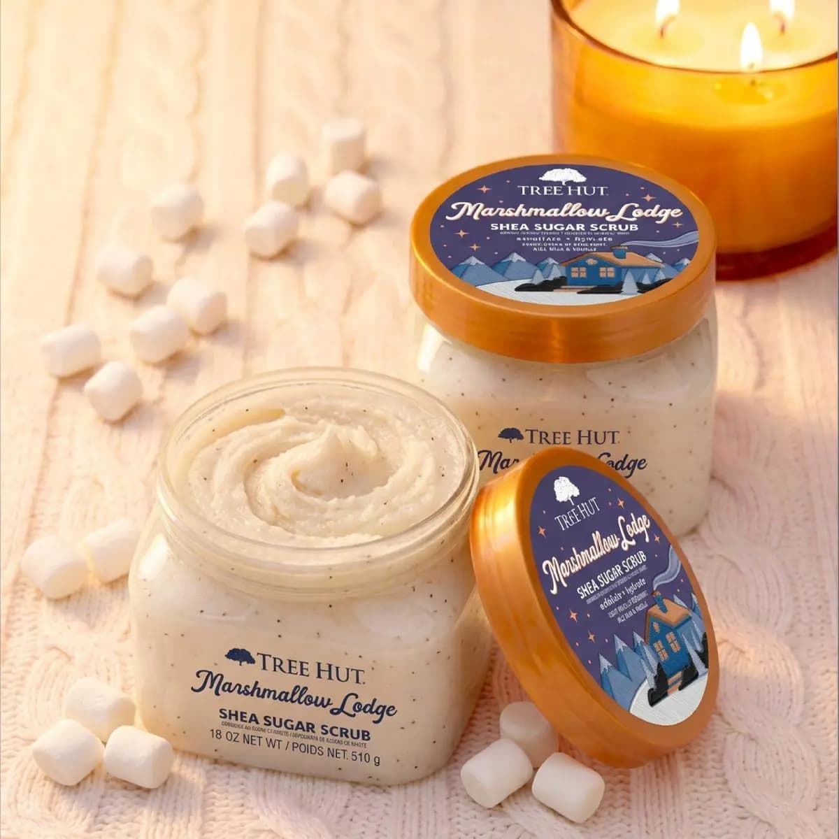 Two Marshmallow Lodge Shea Sugar Scrub jars with mini marshmallows scattered and a warm candle glowing behind