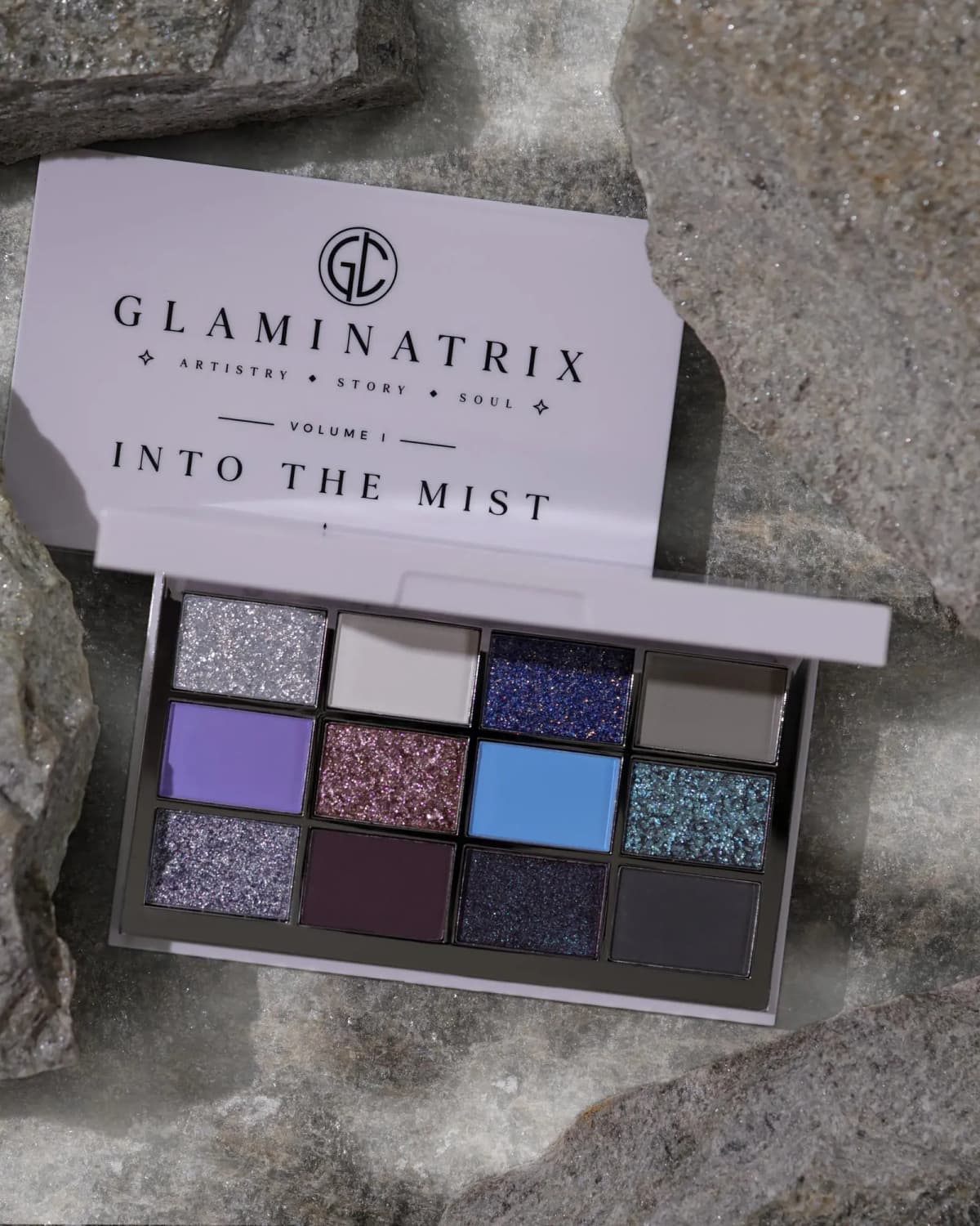 Into the Mist palette open on grey rocks, revealing 12 cool-toned mattes, shimmers and glitters.