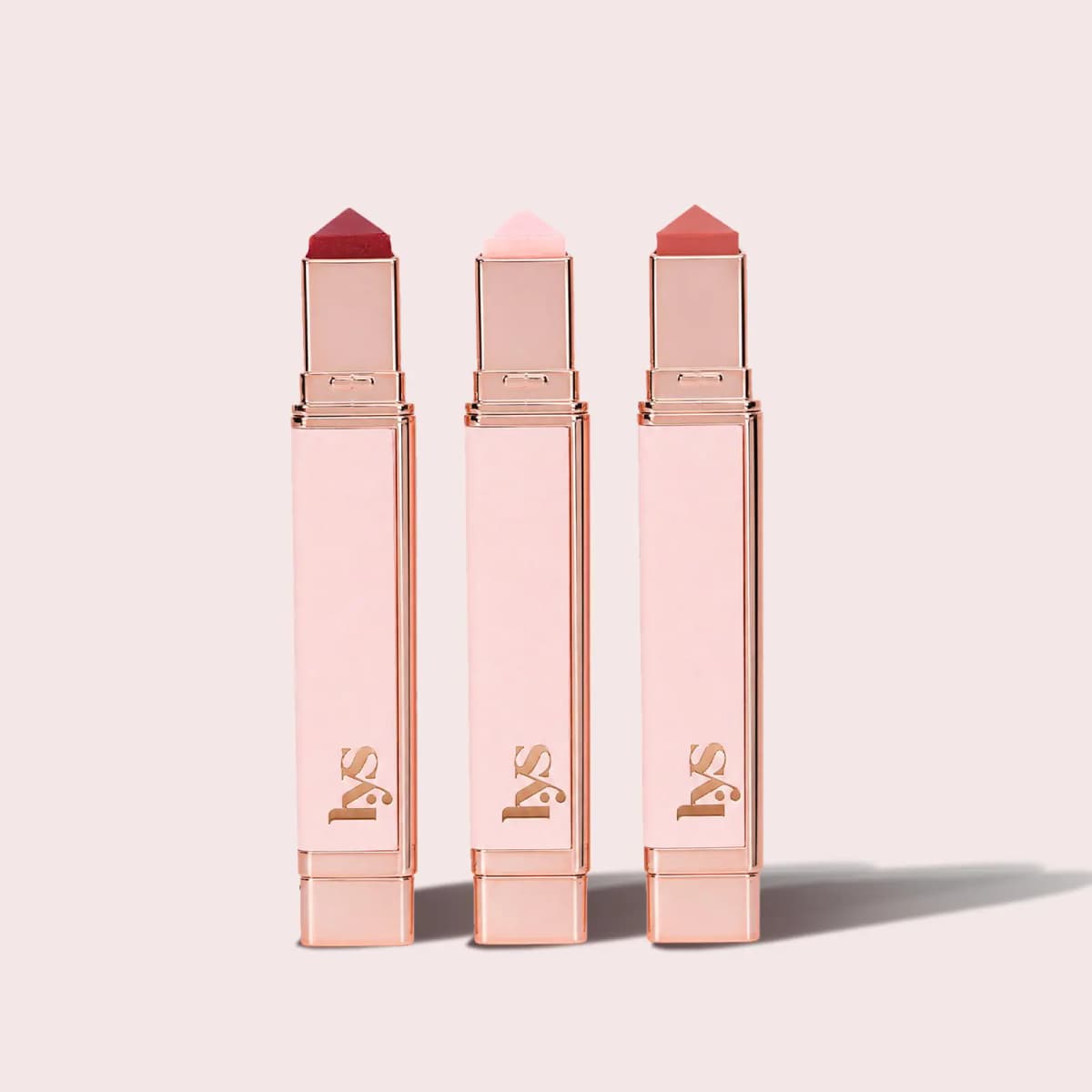 Three rose-gold mini lip balm sticks with pointed bullets in deep red, pale pink and coral on a light pink backdrop