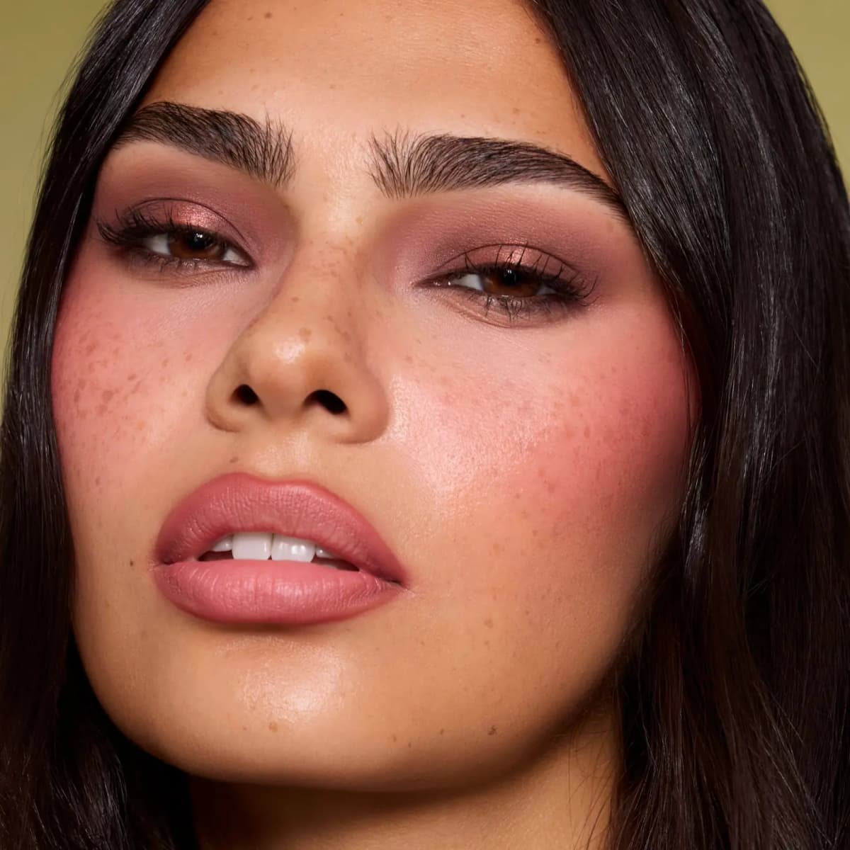 Freckled model with copper and mauve eyeshadow, flushed pink cheeks and matte rosy lips on olive backdrop