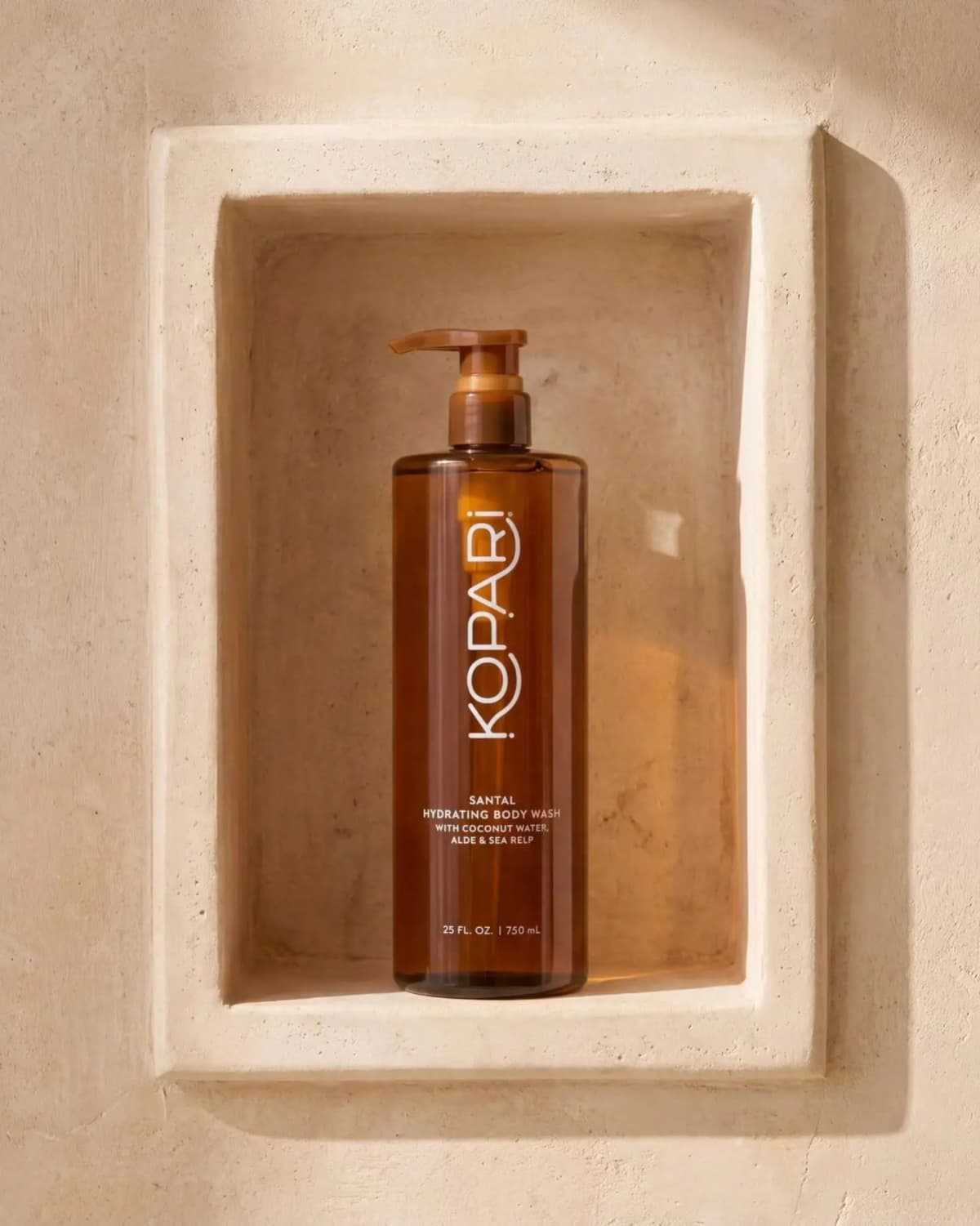 Tall amber Kopari Santal Hydrating Body Wash bottle displayed in a beige stone shower niche with warm sunlight.