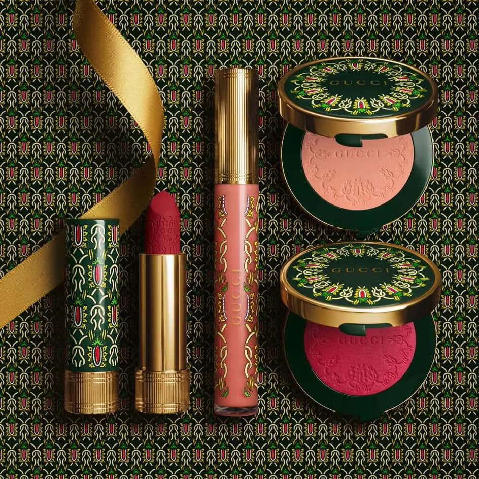 Flat lay of a patterned green lipstick cap, red lipstick, peach gloss and peach and pink blushes with a gold ribbon