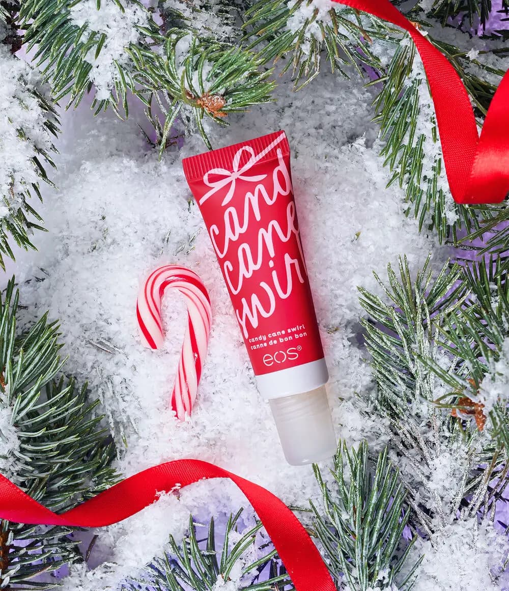 Eos Candy Cane Swirl red tube in snowy pine branches beside a mini candy cane and red satin ribbon