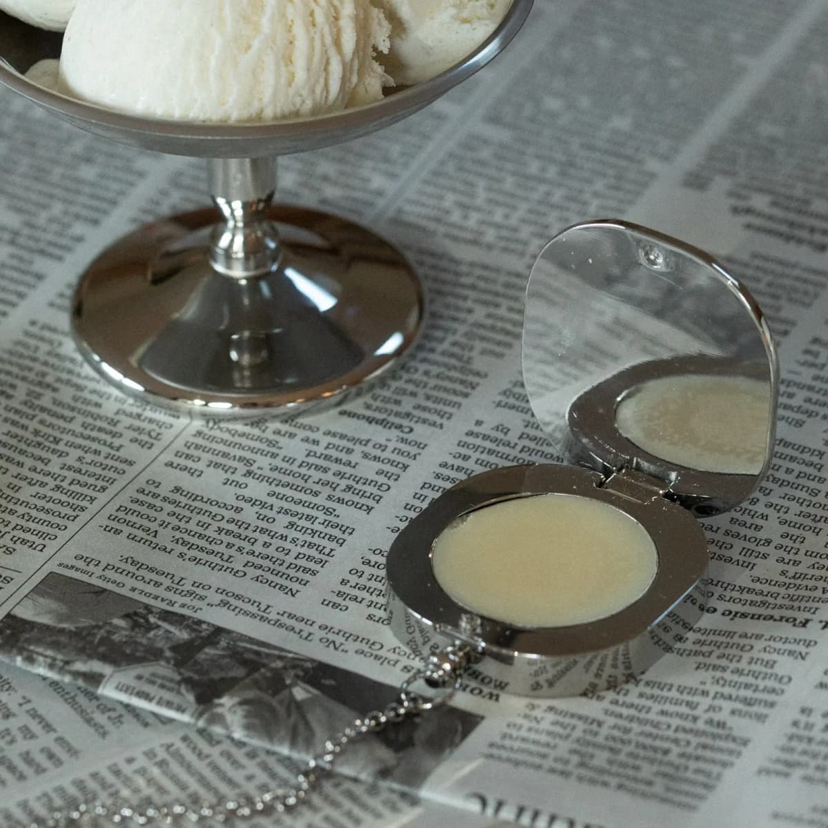 Open mirrored silver compact of pale balm on a chain, set on newspaper beside a dish of vanilla ice cream