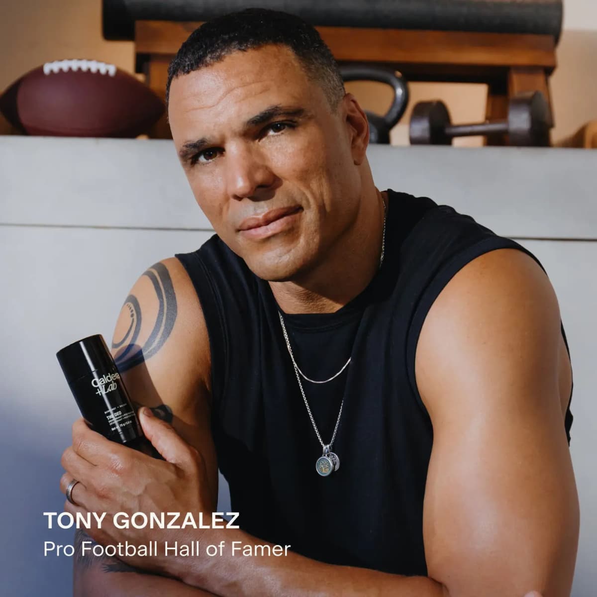 Tony Gonzalez in a black sleeveless top holds the black deodorant stick in a gym with a football and weights