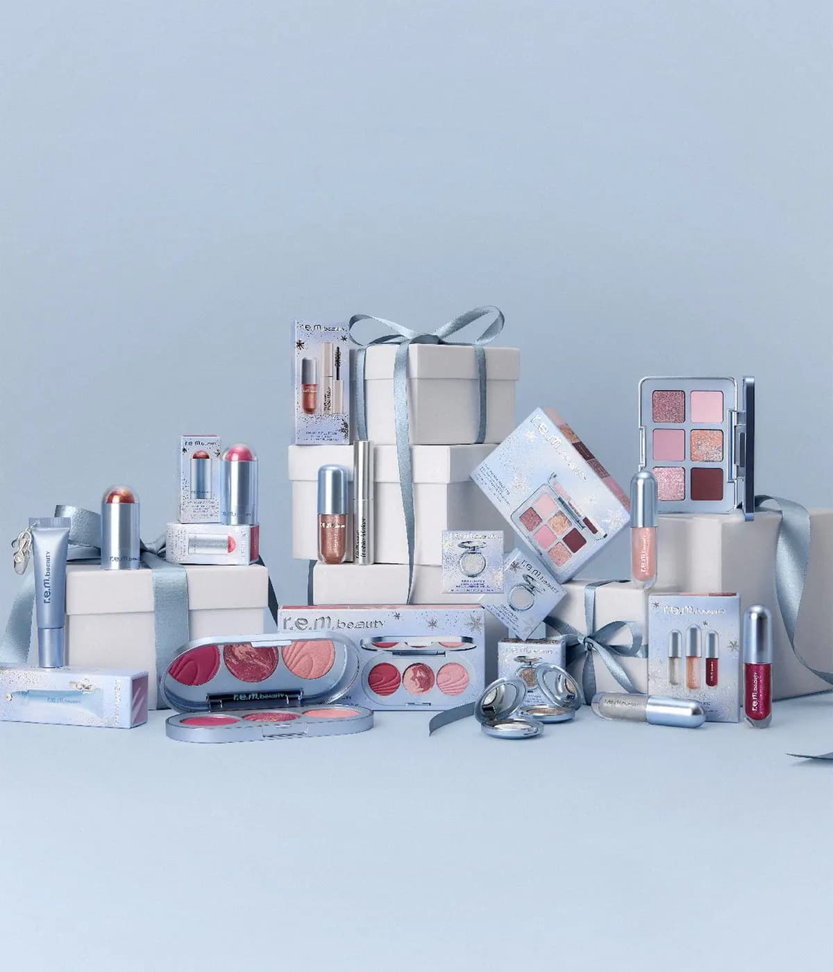R.e.m. beauty holiday makeup arranged around white gift boxes tied with pale blue satin ribbon on a blue backdrop