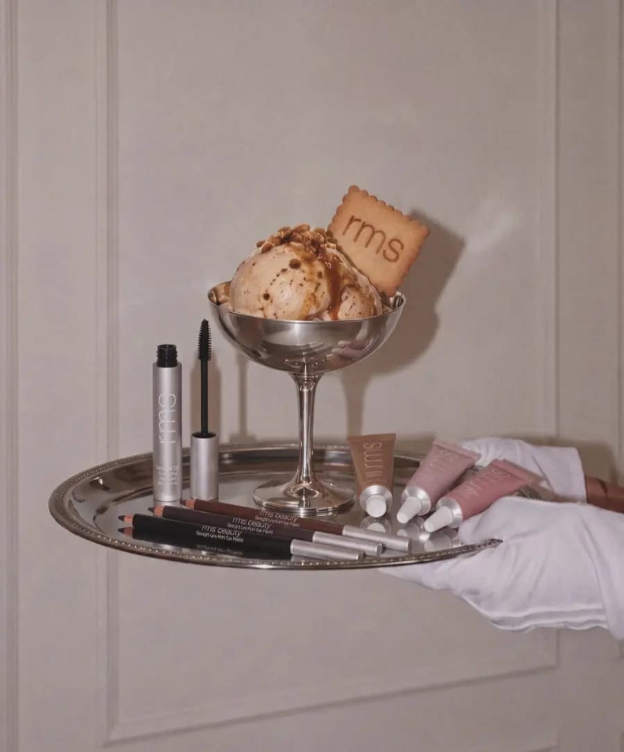 Gloved hand holds a silver tray of mascara, kohl pencils and shadow tubes beside an ice cream sundae with an rms biscuit