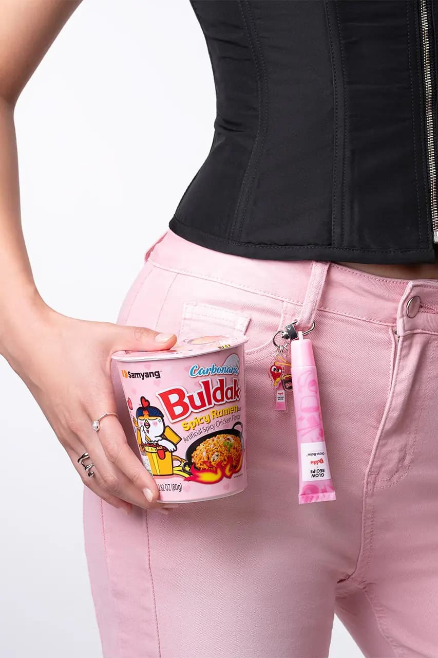 Pink Glass Balm tube clipped to pink jeans beside a Samyang Buldak Carbonara Spicy Ramen cup held in hand