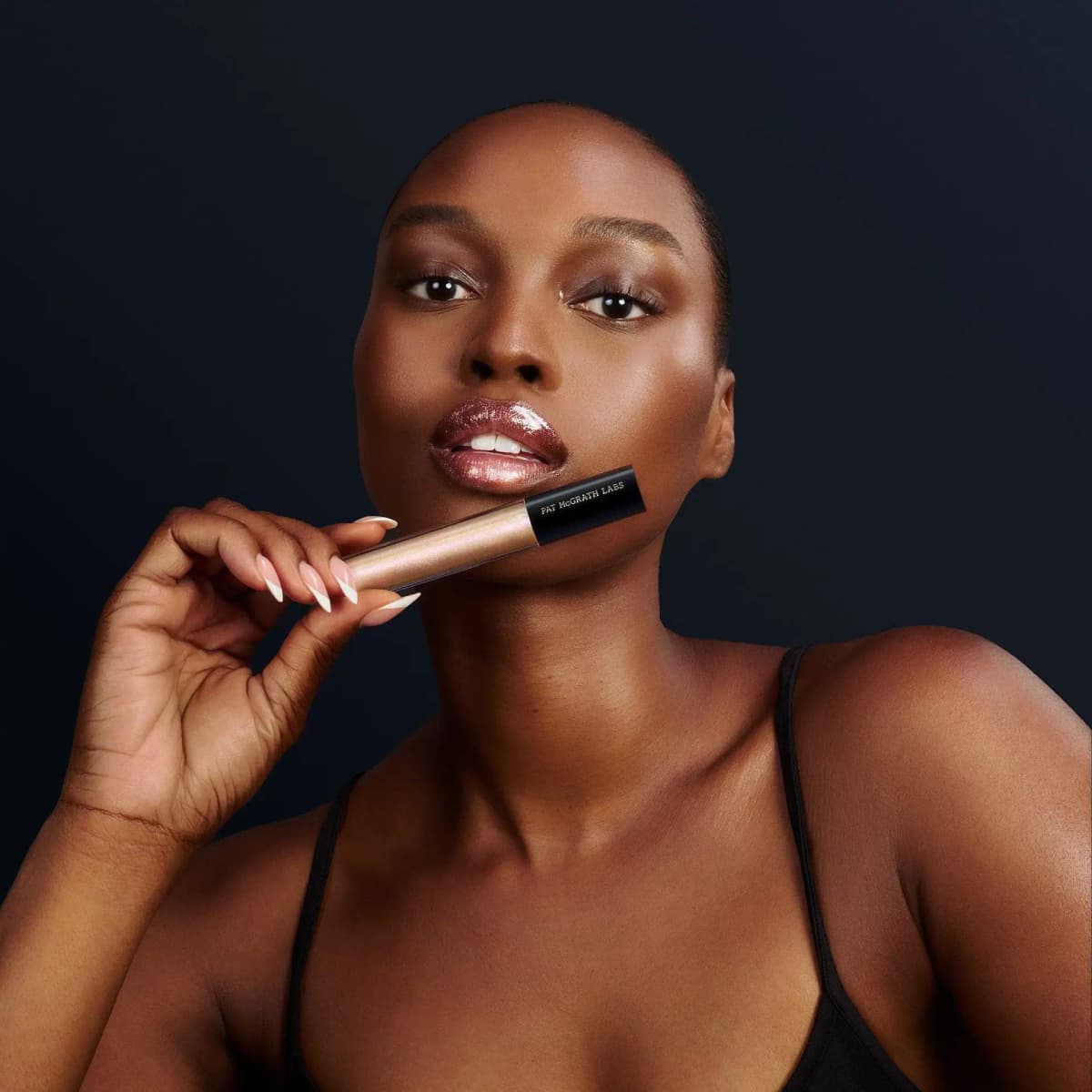 Model with shimmering rosy-bronze glossy lips holds a pink-gold LUST: Gloss tube against a dark backdrop