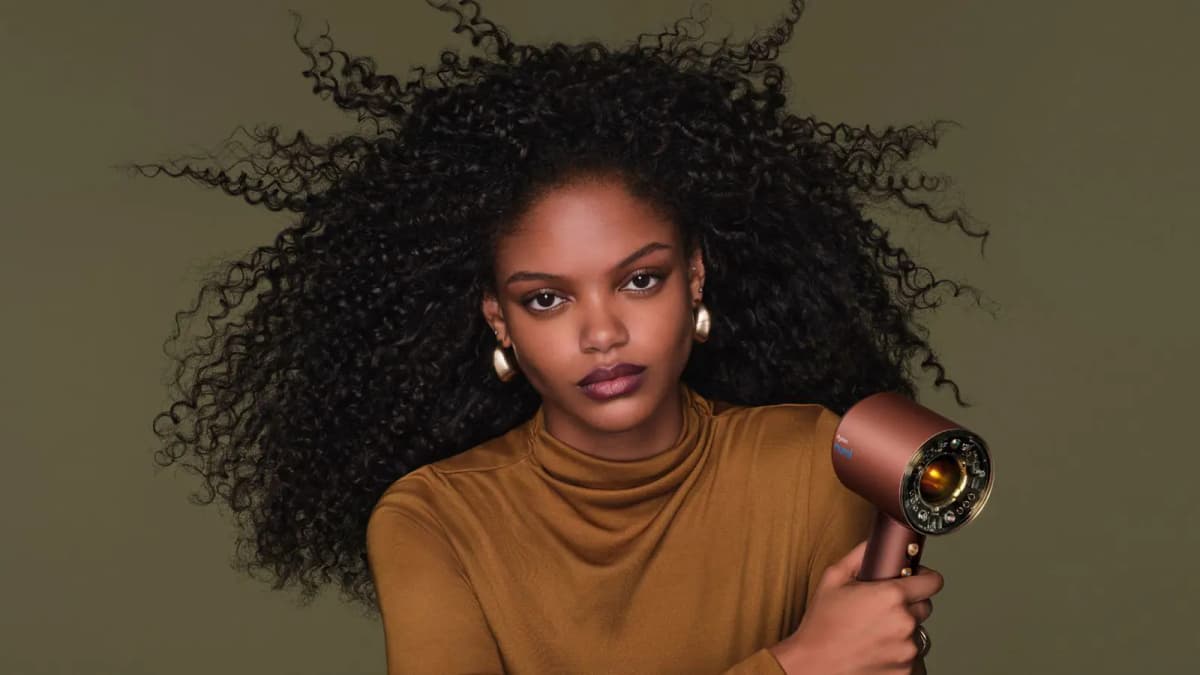Model with voluminous dark curls in a mustard turtleneck holding a copper Dyson Supersonic Nural dryer
