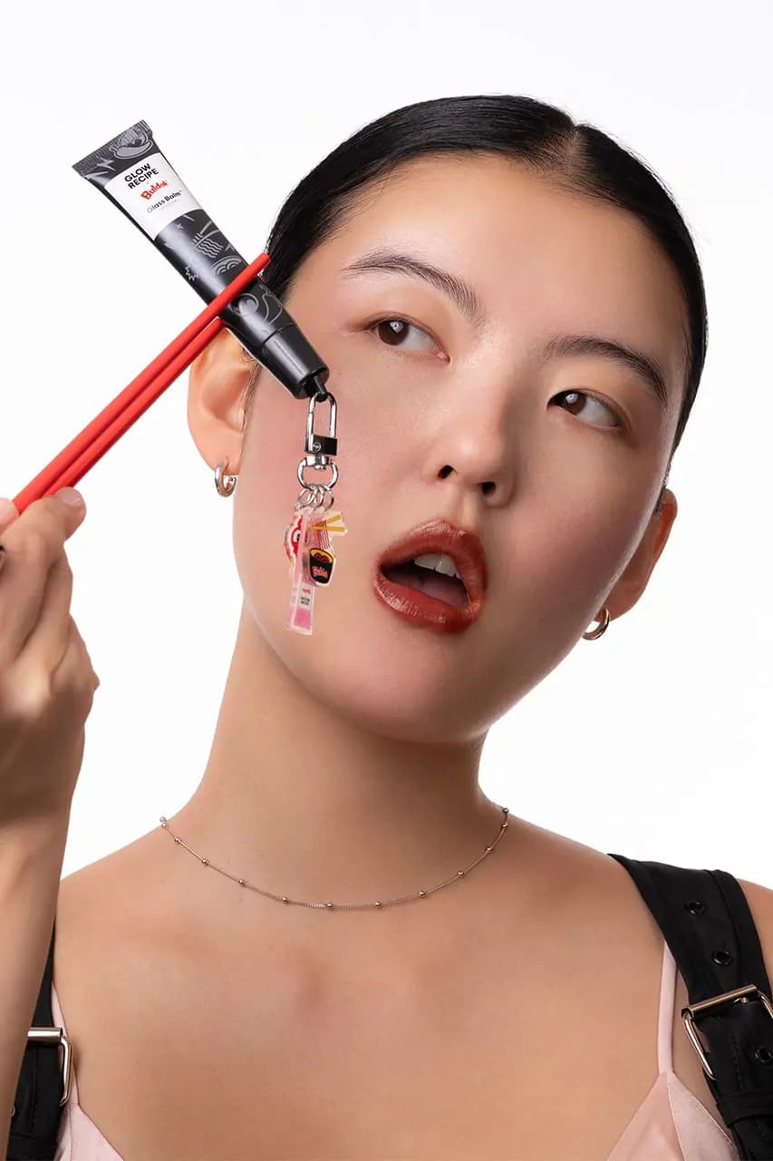Model with glossy brick-red lips holds the black Glass Balm tube with red chopsticks, Peppo charms dangling