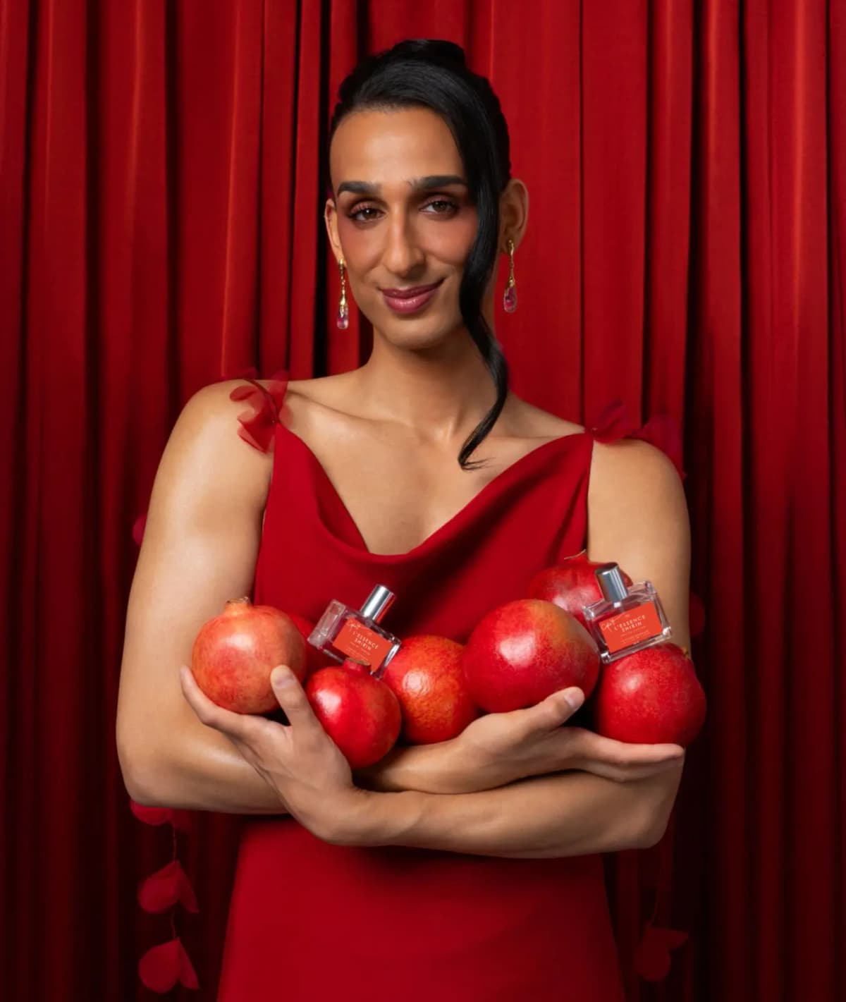 Cyrus Veyssi in a red cowl-neck dress hugging pomegranates and two L'Essence Shirin bottles before a red velvet curtain
