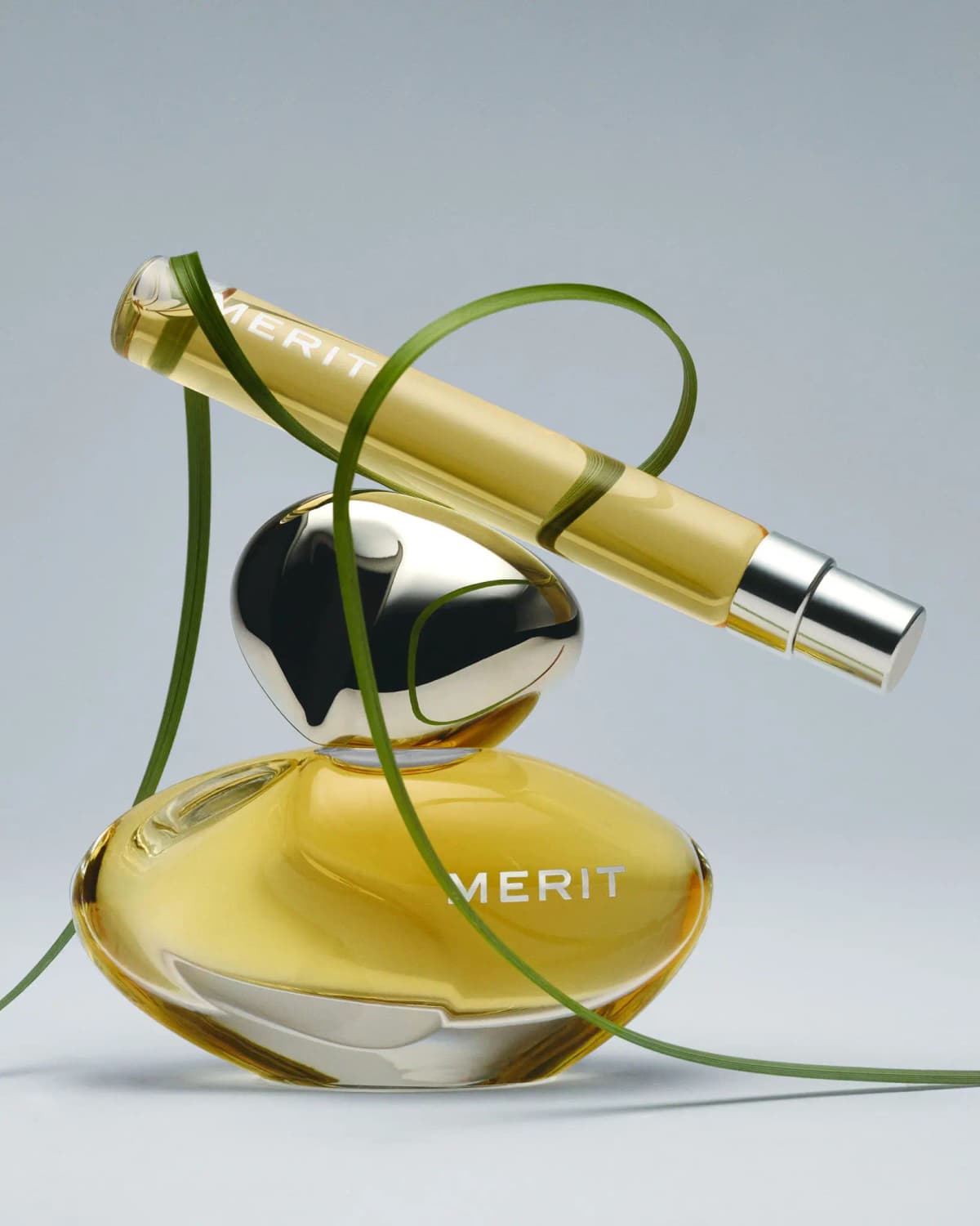 Bottle with silver pebble cap and yellow MERIT travel spray balanced on top, looped with green grass blades on grey