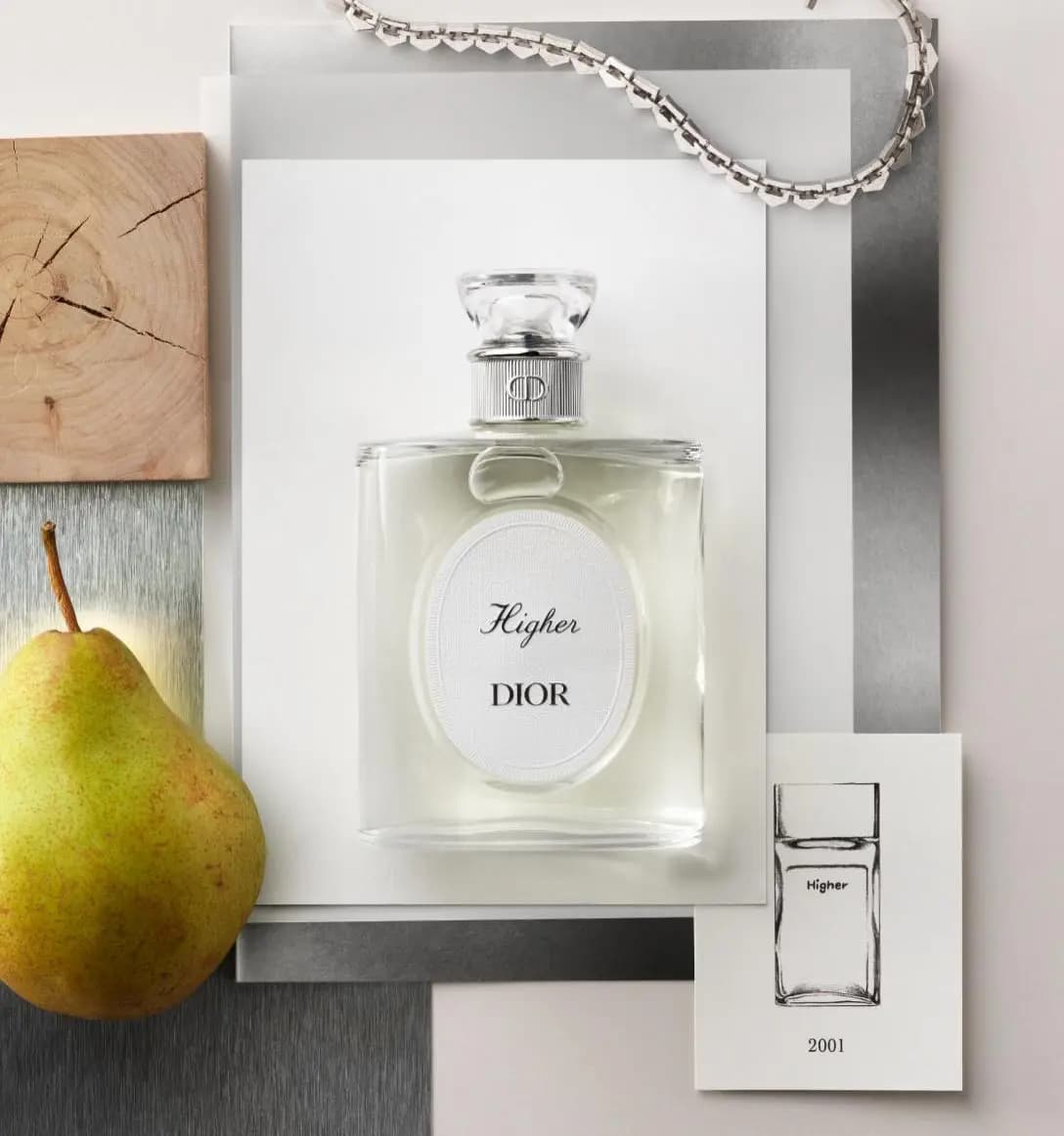 Higher Dior clear fragrance bottle on silver and white paper with a pear, wood block, chain and 2001 sketch card.