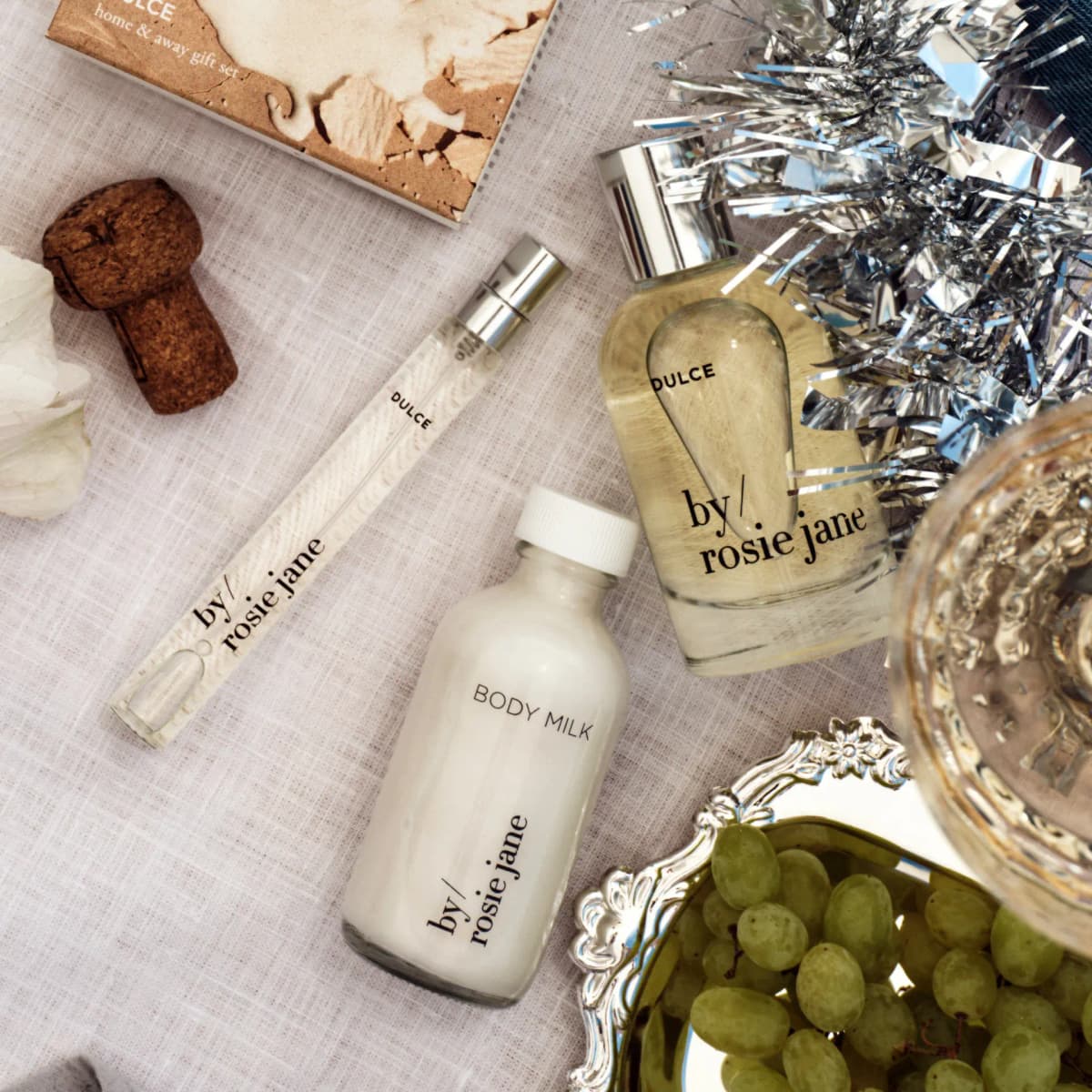 Dulce eau de parfum, travel spray and body milk flat lay on linen with a champagne cork, tinsel and grapes