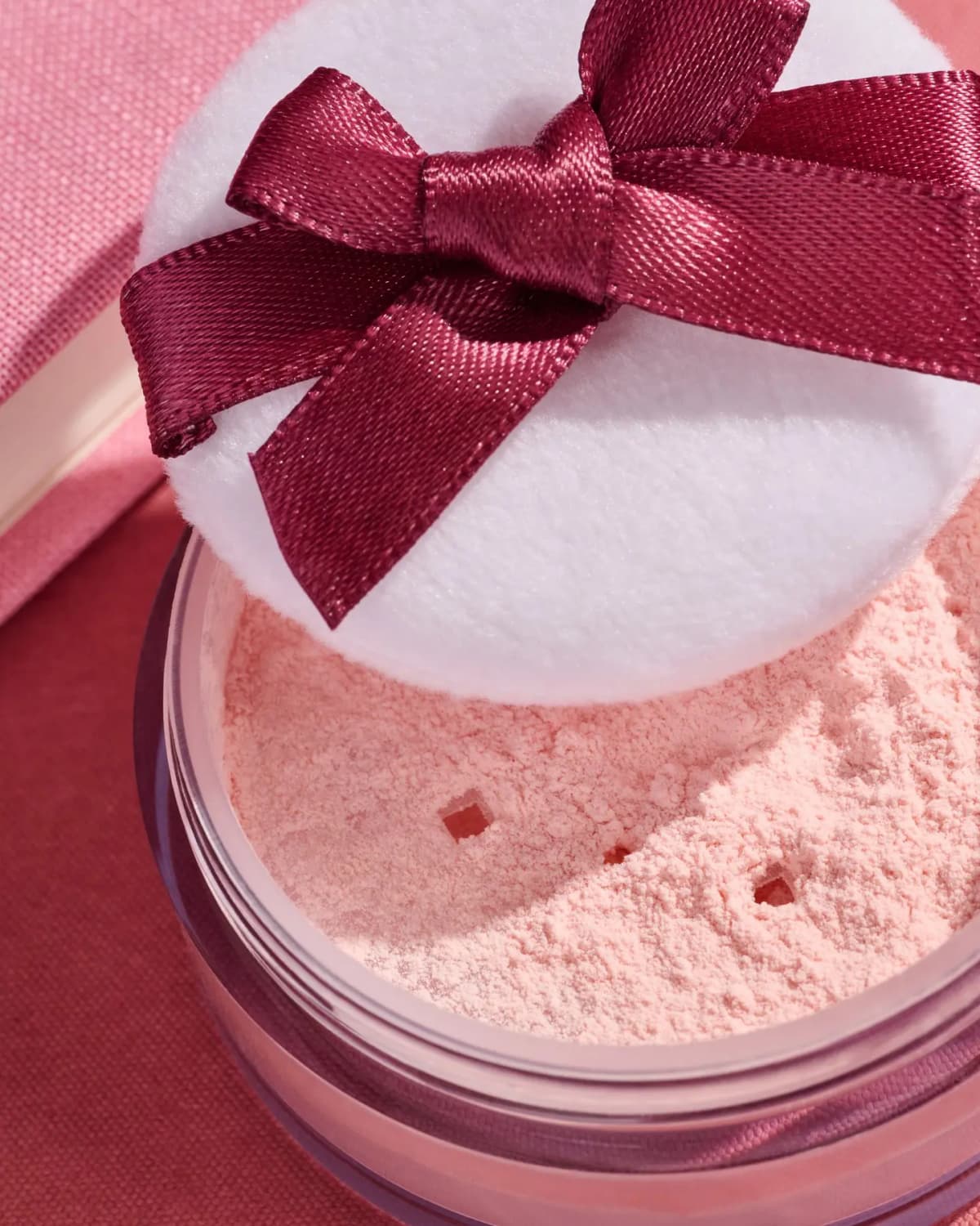 Close-up of peachy-pink loose powder in a jar with a white puff tied with a burgundy satin bow