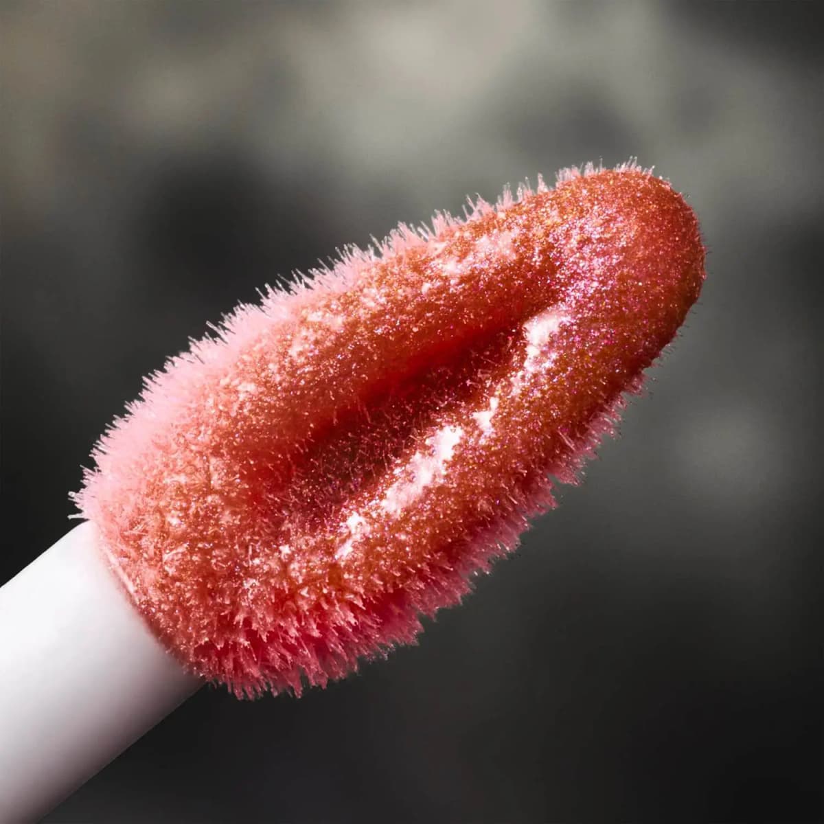Close-up of a fuzzy doe-foot applicator coated in shimmering coral-pink gloss with fine sparkle