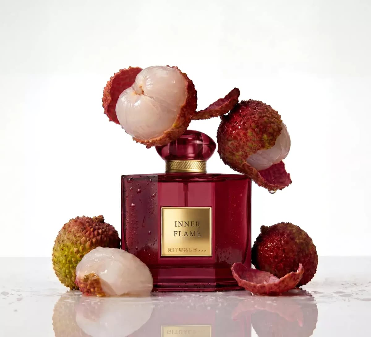 Ruby-red square Rituals Inner Flame bottle with gold label surrounded by whole and peeled lychees on a wet surface