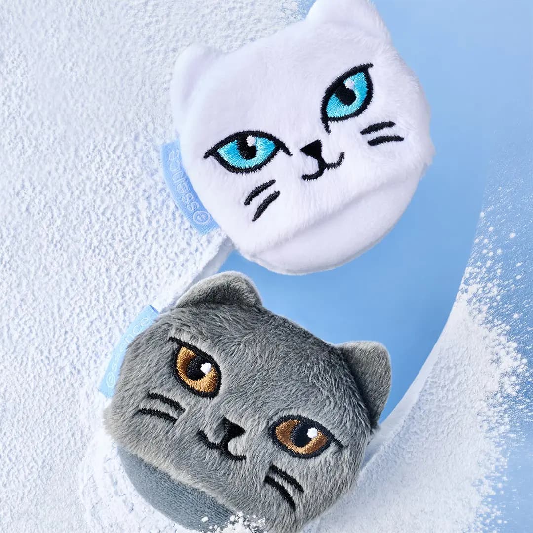 Fluffy white and grey cat-face powder puffs with embroidered eyes resting in loose white powder