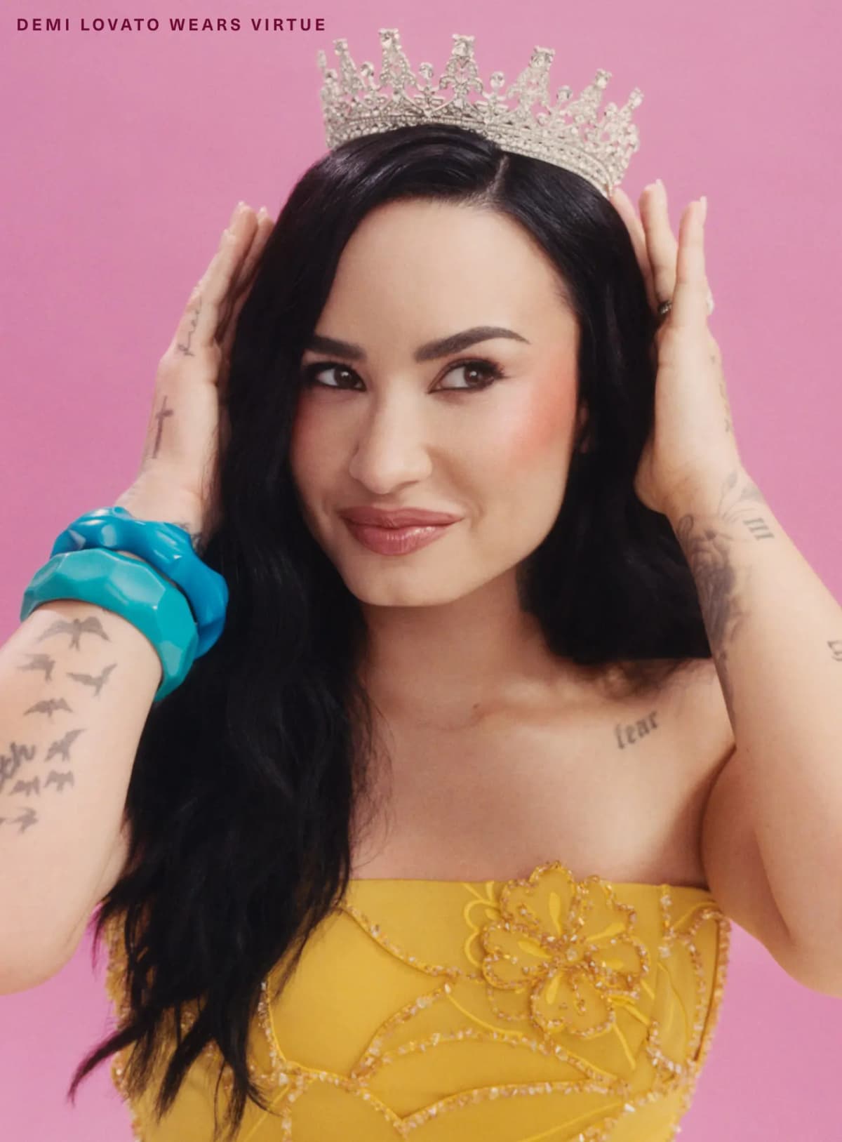 Demi Lovato wearing Virtue blush, adjusting a crystal crown, in a yellow beaded dress and blue bangles on pink