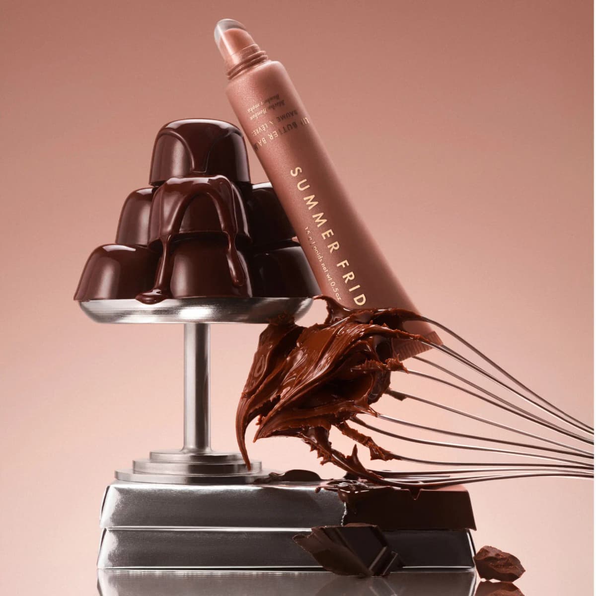 Mocha Bonbon Lip Butter Balm leaning on a stand of glossy chocolates beside a chocolate-coated whisk