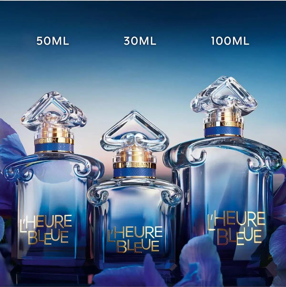 Three L'Heure Bleue bottles labelled 50ml, 30ml and 100ml lined up against a dusky blue sky with iris petals.