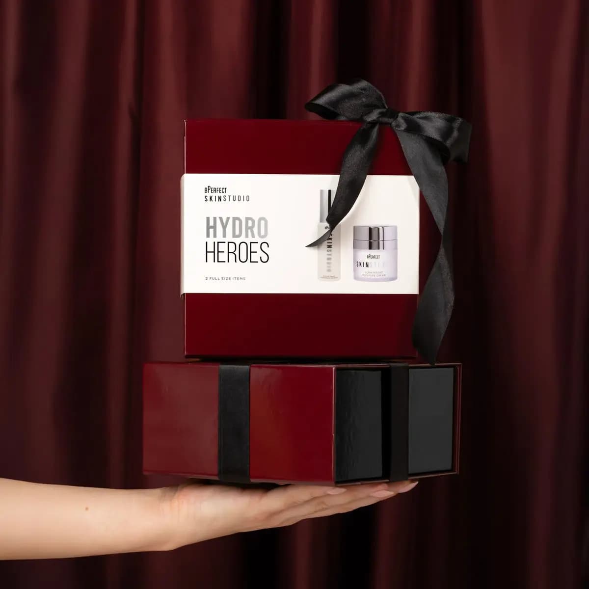 Hand holding a BPerfect Skin Studio Hydro Heroes box tied with a black satin bow atop a burgundy gift box
