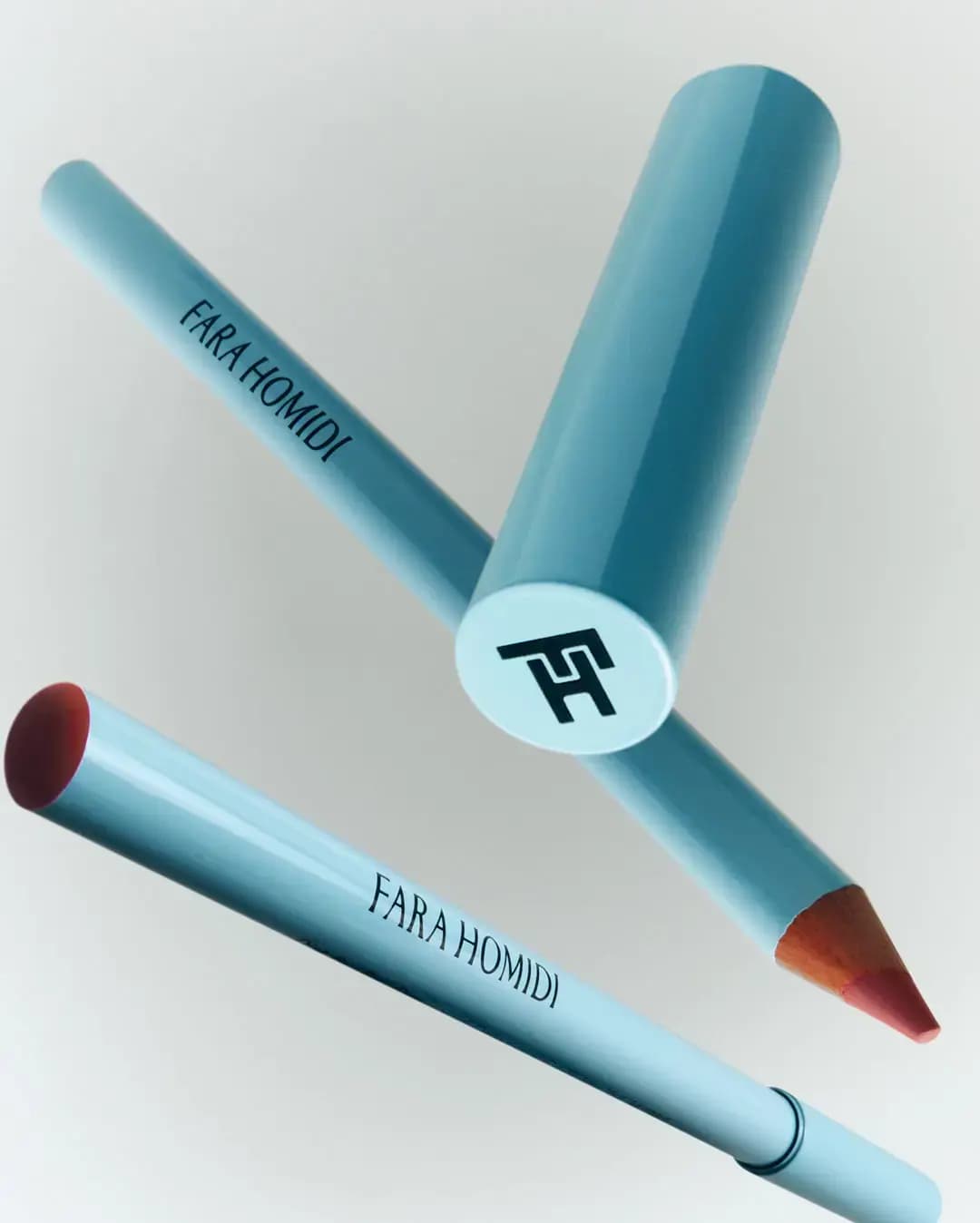 Two light-blue Fara Homidi lip pencils floating, one sharpened to a rosy tip, beside a cap with the FH monogram