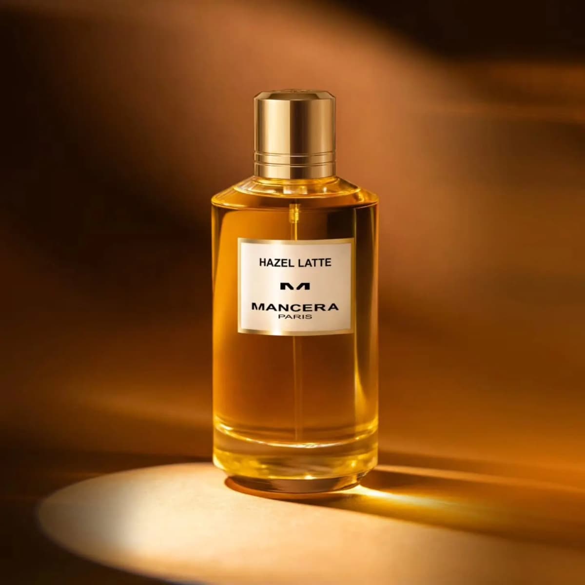 Mancera Hazel Latte perfume bottle with amber liquid and a gold cap, lit by a warm spotlight against a bronze backdrop