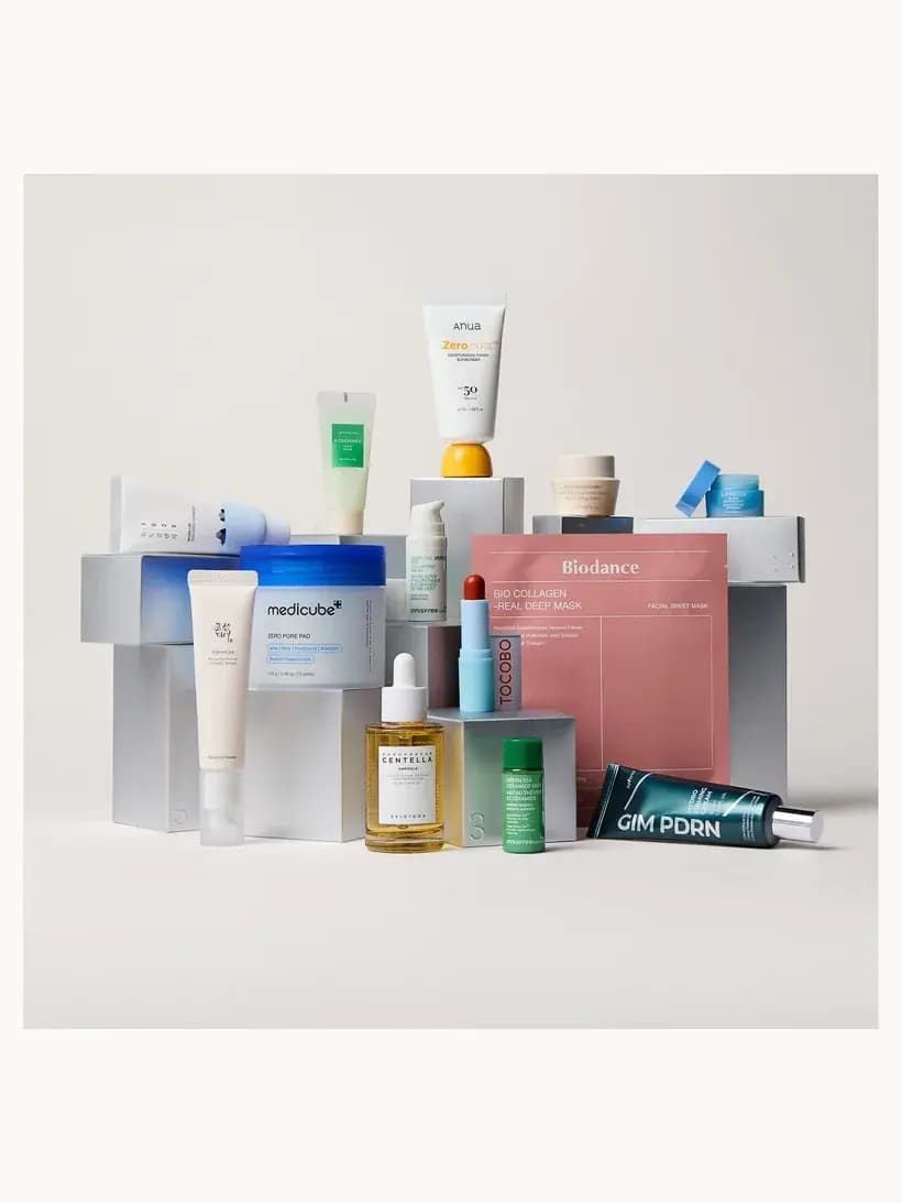 Twelve K-beauty products arranged on grey pedestals, including Anua sunscreen, Medicube pads, Biodance mask and Tocobo lip