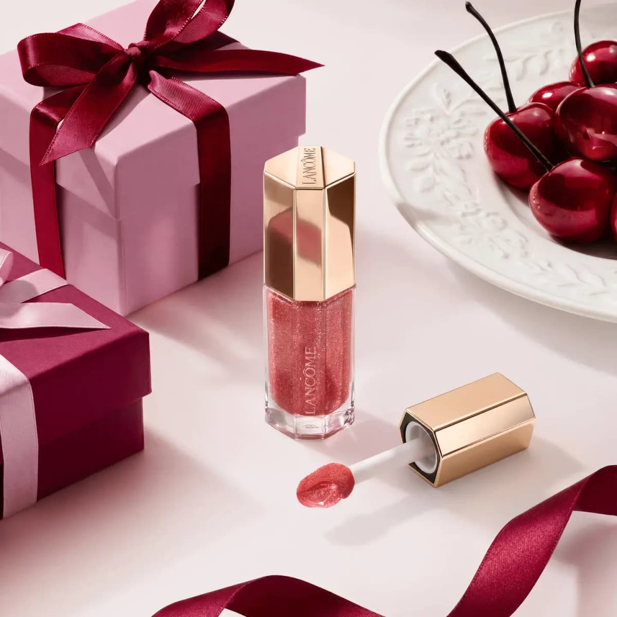 Shimmering berry lip oil gloss with gold cap beside pink and burgundy gift boxes, satin ribbon and a plate of cherries