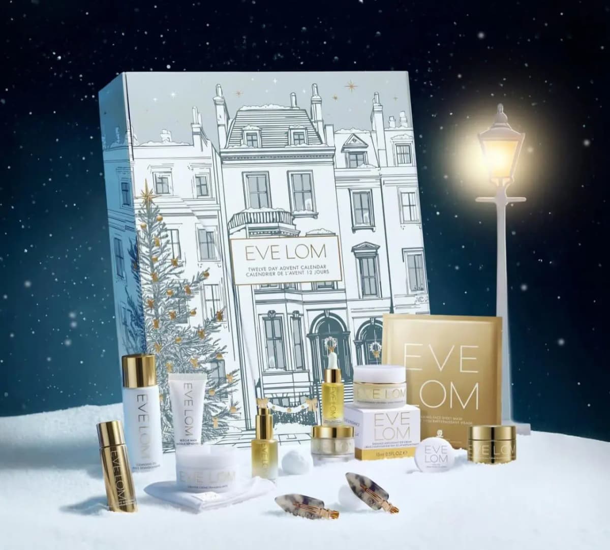EVE LOM Twelve Day Advent Calendar with gold and white skincare, hair clips and sheet mask in snow beside a glowing lamppost