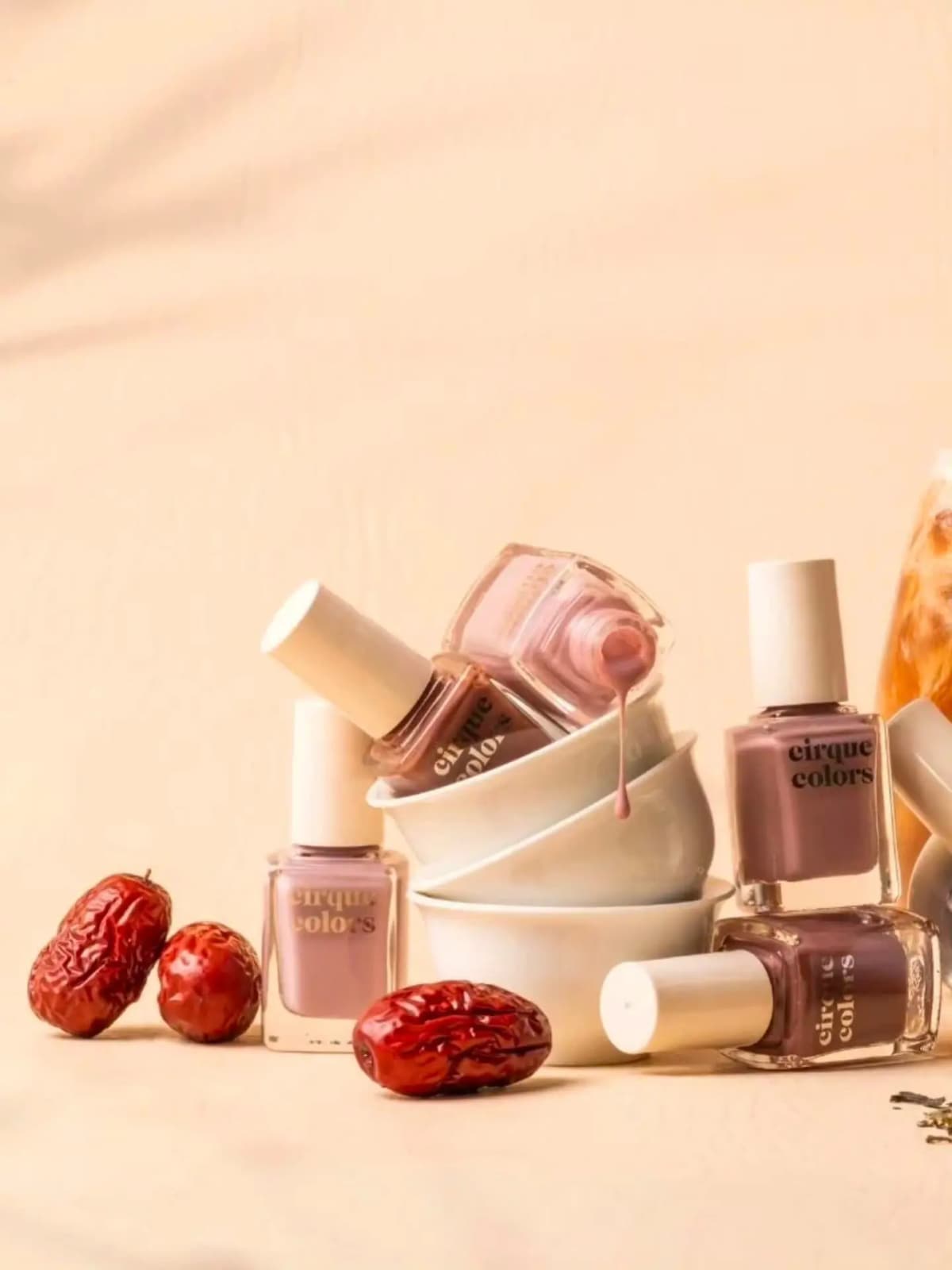 Dusty pink polish drips from a tipped bottle into stacked white bowls beside mauve bottles and dried jujubes