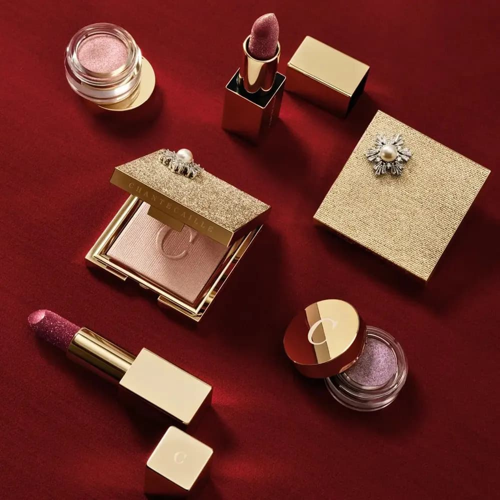Chantecaille Bijoux d'Or Holiday 2026 Collection flat lay with the Cheek Light compact, Eye Frost jars and Lip Cristal lipsticks on red fabric
