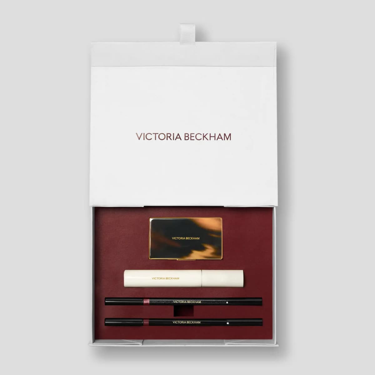 Open white box lined in burgundy holding a tortoiseshell-and-gold compact, cream mascara and two black liners