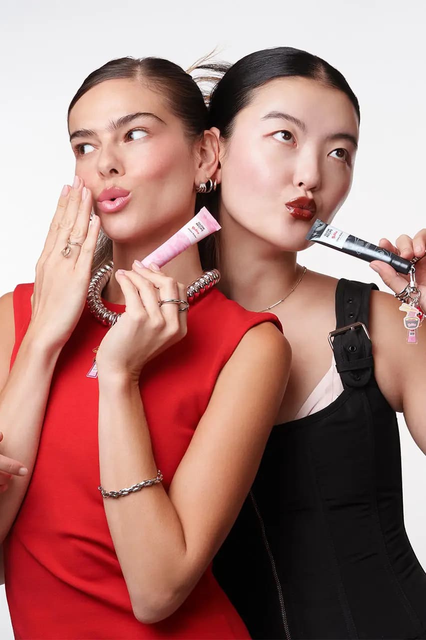 Two models pout, one with sheer pink lips holding the pink tube, the other with deep red lips holding the black tube