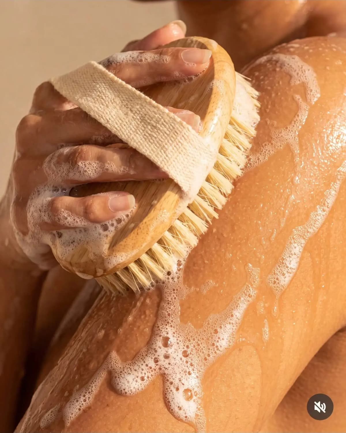 Close-up of soapy hands using a wooden bristle body brush on a foam-covered shoulder and arm.