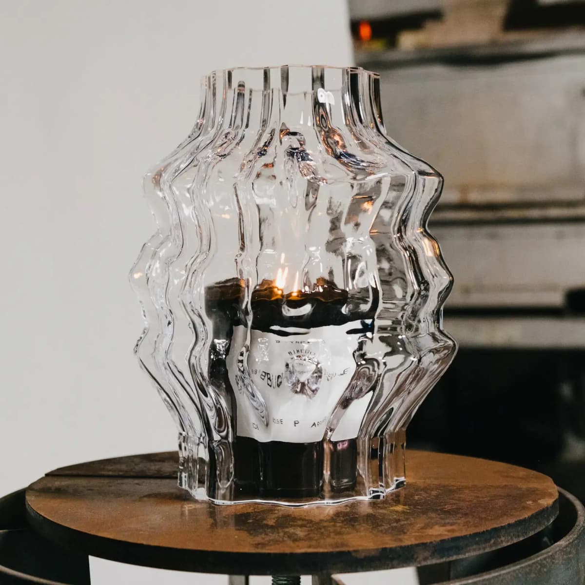 Lit Bibliothèque candle glowing through a rippled glass hurricane on a rusted metal stool