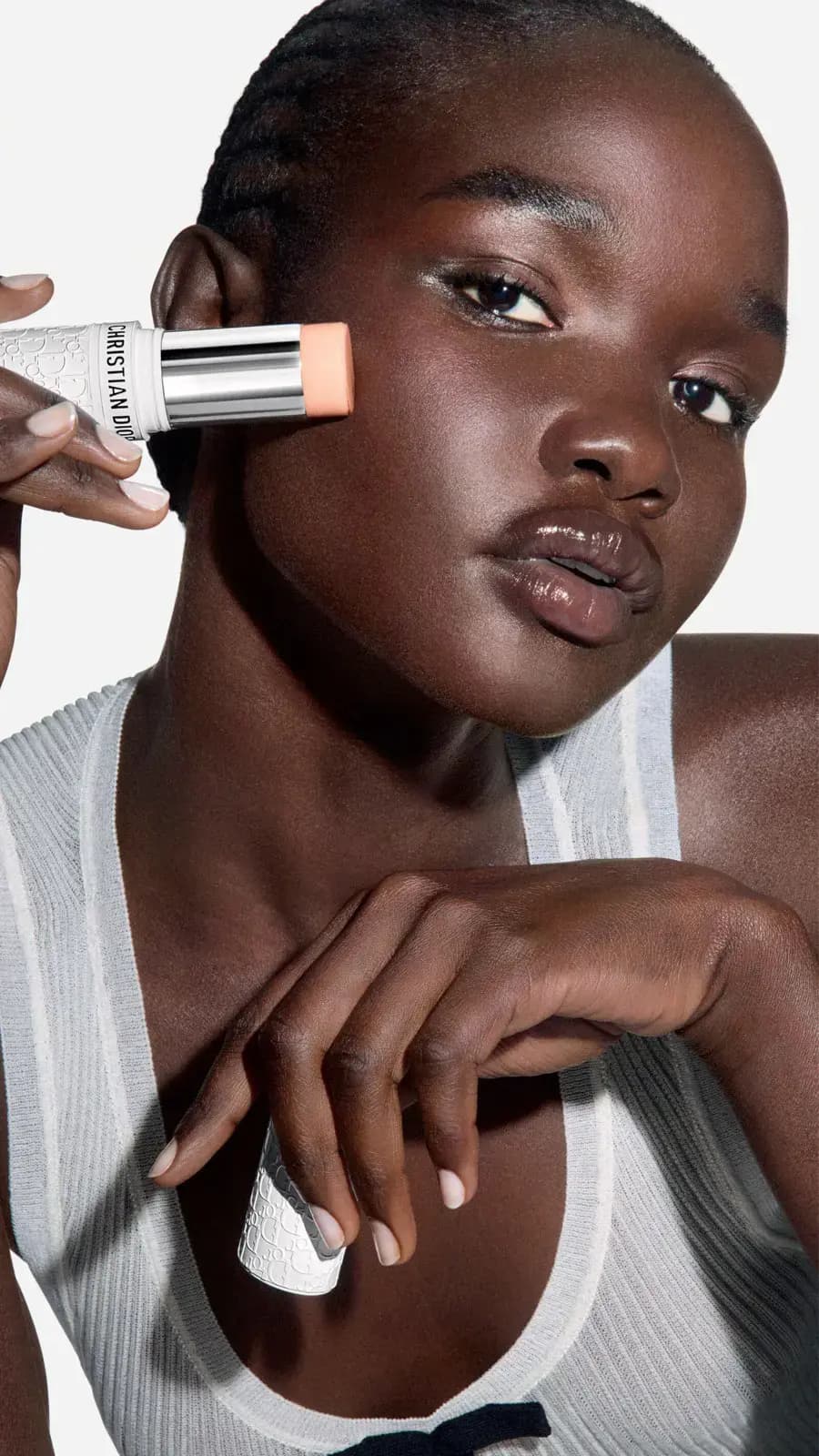 Model with deep skin tone and glossy lips presses the stick to her cheekbone, cap in her other hand