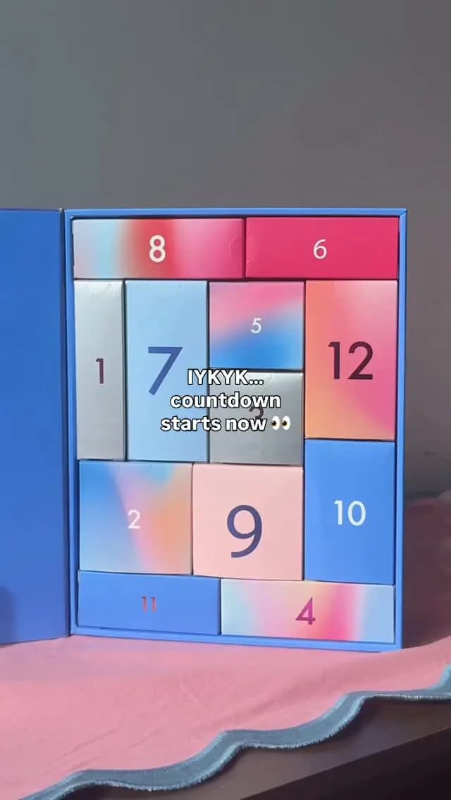 Hands-on shot of the open calendar's numbered doors on a pink cloth, with 'IYKYK… countdown starts now' text